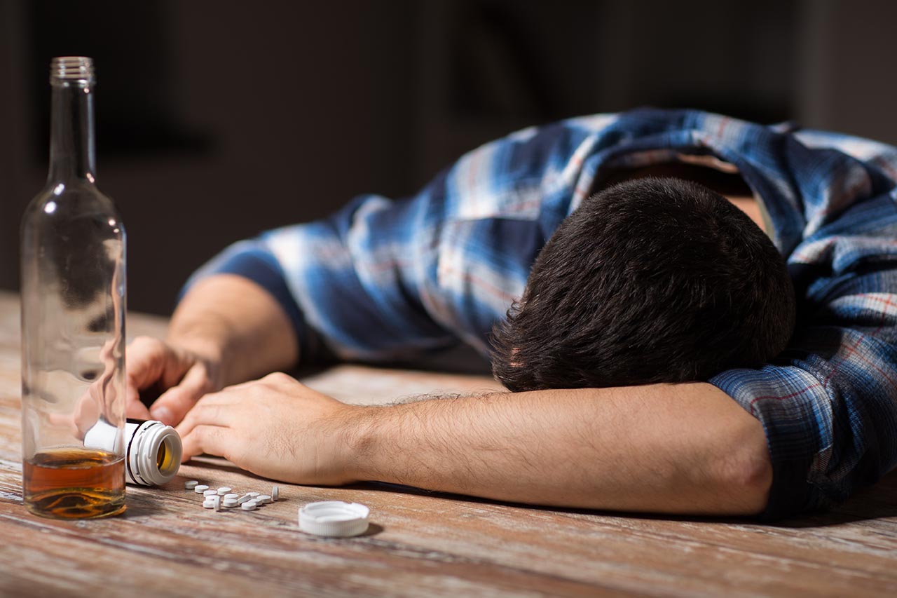 man with his head on the table next to an empty bottle of alcohol and pills