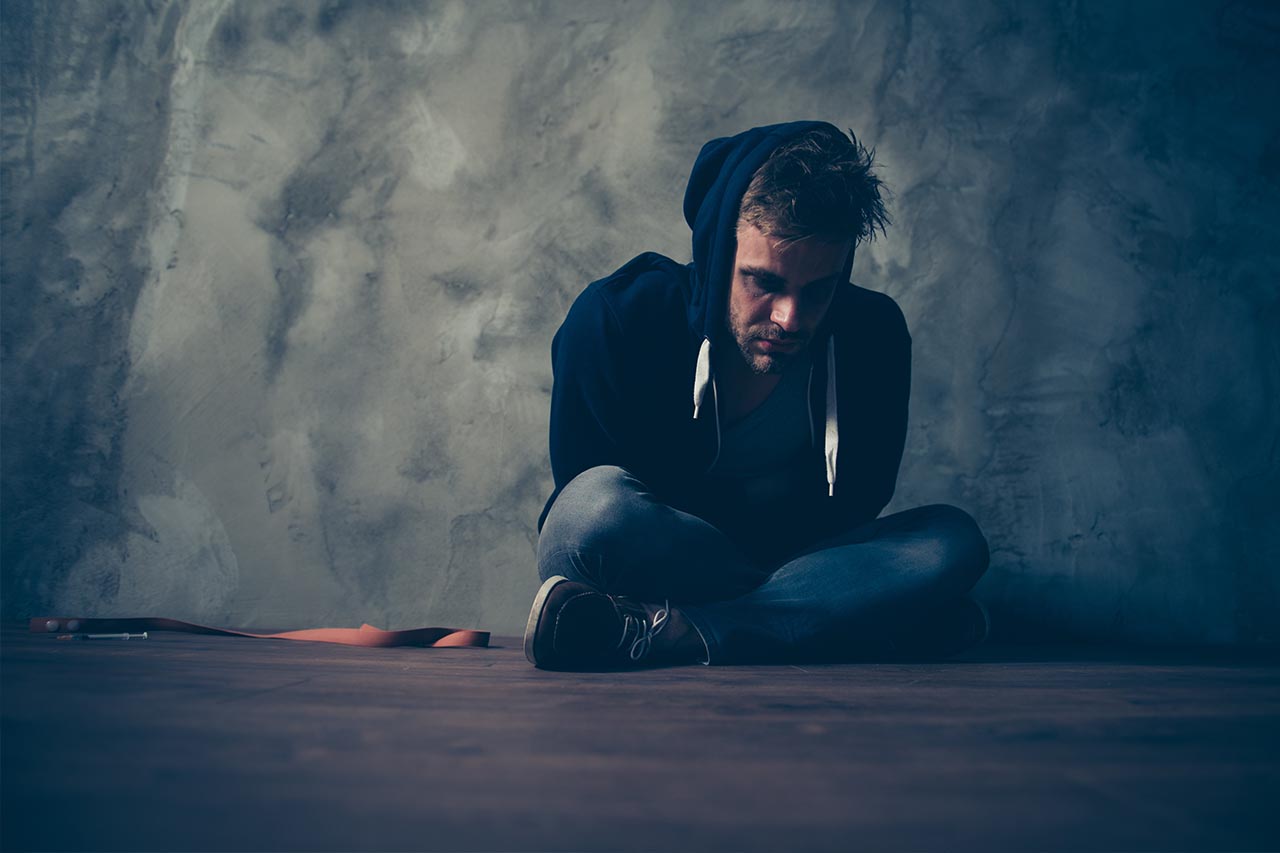 Man in a black hoodie singing on the floor next to a syringe and rubber strap